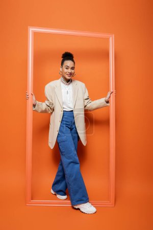 Téléchargez les photos : Attrayant jeune modèle de mode en blazer beige avec un maquillage vif debout et tenant cadre orange - en image libre de droit