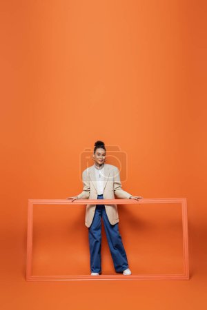 modelo afroamericano sonriente en chaqueta beige y pantalones azules sosteniendo marco sobre fondo naranja