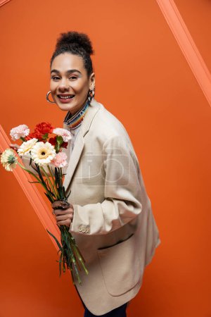 cheerful trendy fashion model in chic outfit with sleek bun grasping orange framework and flowers