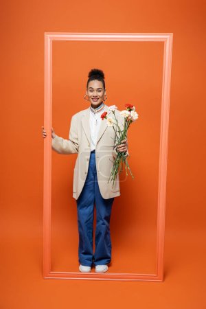 smiling trendy african american woman in fashionable outfit holding orange frame and flower bouquet