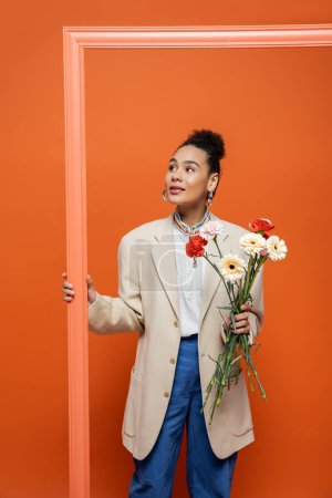 modèle de mode souriant en tenue tendance avec accessoires tenant cadre orange et bouquet de fleurs