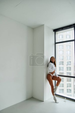 sexy woman with pierced belly in long sleeve shirt and panties standing near window at home