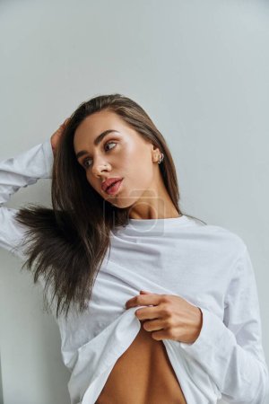 Photo for Slim woman with long hair adjusting long sleeve shirt and looking away near white wall - Royalty Free Image
