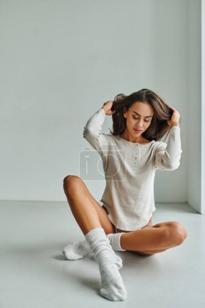 Foto de Mujer joven sonriente en calcetines y camisa de manga larga sentada en el suelo en casa y ajustando el pelo largo - Imagen libre de derechos