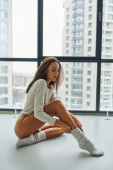 beautiful young woman in long sleeve shirt sitting on floor and wearing socks near panoramic window Poster #674383010