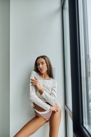 brünette junge Frau in langärmeligem Hemd und Höschen, die in der Nähe des Panoramafensters steht, sexy