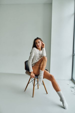 nachdenkliche und sexy Frau in langärmeligem Hemd und Socken zu Hause auf Stuhl sitzend, Weiblichkeit