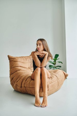 Nachdenkliche junge Frau mit nackten Füßen sitzt auf bequemen Sitzsäcken in Pflanzennähe, Wochenendstimmung