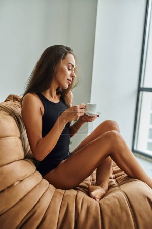 hübsche junge Frau mit brünetten langen Haaren, die eine Tasse Morgenkaffee in der Hand hält und auf einem Bohnensackstuhl sitzt