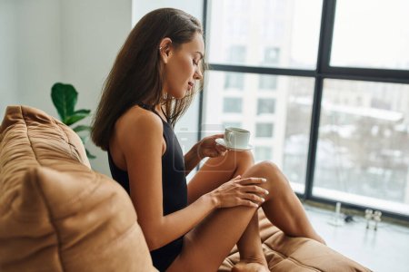 Téléchargez les photos : Vue latérale, jeune femme aux cheveux bruns tenant une tasse de café du matin, assise sur une chaise de sac de haricots - en image libre de droit