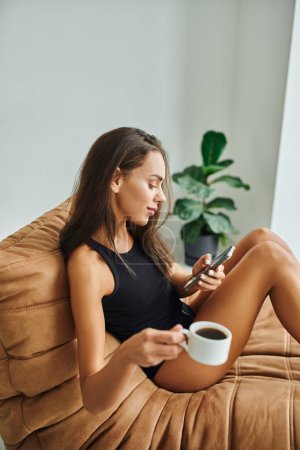 glückliche Frau mit Smartphone und einer Tasse schwarzen Kaffees, die zu Hause auf einem Sitzsack-Stuhl sitzt