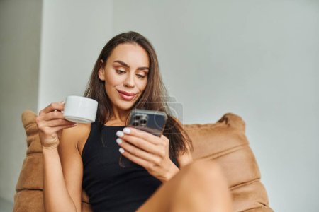 Téléchargez les photos : Femme positive en utilisant un smartphone et tenant une tasse de café, assis sur une chaise de sac de haricots à la maison - en image libre de droit