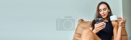 Téléchargez les photos : Femme positive utilisant smartphone et tenant une tasse de café, assis sur une chaise de sac de haricot à la maison, bannière - en image libre de droit