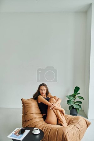 Foto de Atractiva mujer descalza sentada en la silla de la bolsa de frijol cerca de la mesa de café con taza de café negro - Imagen libre de derechos