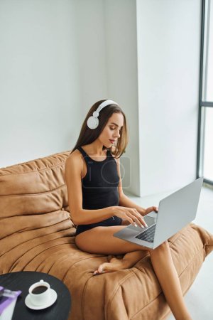 descalzo freelancer en auriculares inalámbricos utilizando el ordenador portátil y sentado en la silla de la bolsa de frijoles, mujer bonita