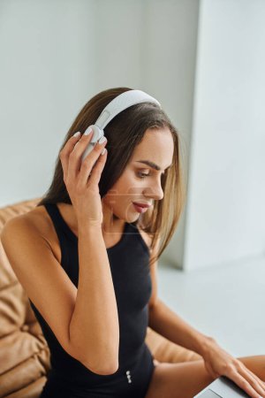 Téléchargez les photos : Belle et brune femme aux cheveux longs écoutant de la musique dans des écouteurs sans fil à la maison - en image libre de droit