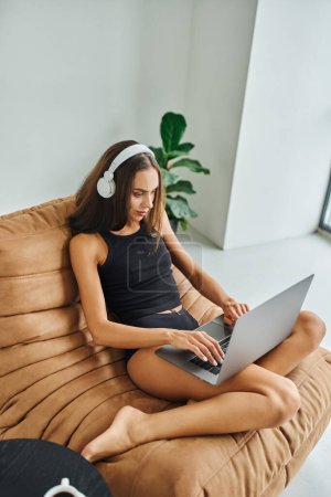 hermosa freelancer en auriculares inalámbricos utilizando el ordenador portátil y sentado en la silla de la bolsa de frijol, mujer joven