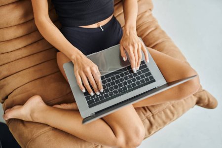 vista recortada de freelancer en camiseta sin mangas negro usando el ordenador portátil y sentado en la silla de la bolsa de frijol, mujer joven