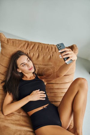hermosa mujer en camiseta sin mangas y bragas descansando en la silla de la bolsa de frijoles y tomando selfie en el teléfono inteligente