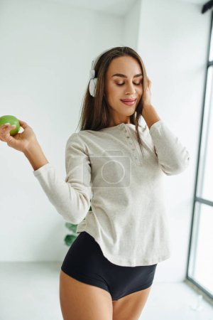 joyful woman in long sleeve and panties listening music in headphones and holding apple