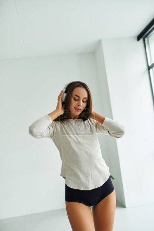 Téléchargez les photos : Heureux brunette femme en manches longues et culotte marche tout en écoutant de la musique dans les écouteurs - en image libre de droit