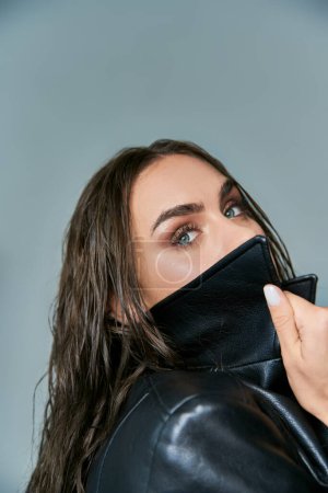 alluring woman with wet hair and blue eyes adjusting collar of black leather coat on grey backdrop