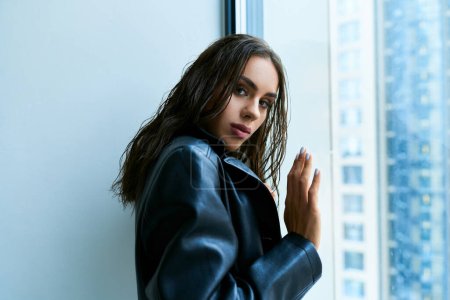 sexy Frau mit brünetten nassen Haaren posiert in schwarzem Ledermantel und berührt Fenster zu Hause