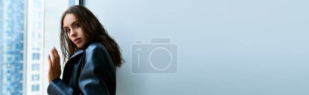 Foto de Mujer sexy con pelo mojado morena posando en abrigo de cuero negro y tocando la ventana en casa, pancarta - Imagen libre de derechos