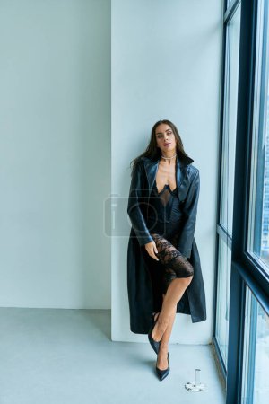 passionate woman with brunette wet hair posing in black leather coat and lace underwear near window