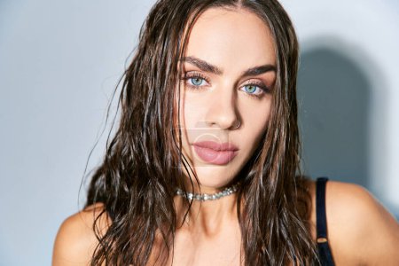 portrait of beautiful woman with brunette and wet hair looking at camera on grey backdrop, blue eyes