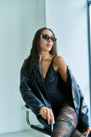 seductive woman in sunglasses, black lace underwear and leather coat sitting on chair, grey backdrop