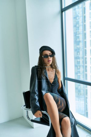 modèle séduisant en lunettes de soleil, béret, sous-vêtements en dentelle et manteau en cuir assis sur la chaise près de la fenêtre