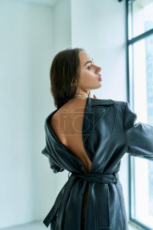 vista trasera de la mujer sexy en abrigo de cuero negro con la espalda abierta de pie cerca de la ventana, elegante y caliente