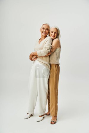 glücklich tätowierte Frau umarmt stilvolle Freundin auf grau, anmutige Alterung der älteren Modelle