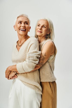 señora tatuada madura abrazando alegre y elegante amiga en gris, concepto de envejecimiento de moda
