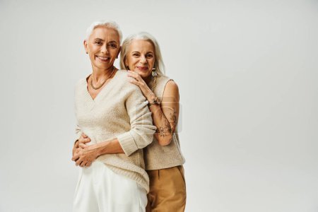 cheerful senior women in fashionable casual attire looking at camera on grey, timeless beauty