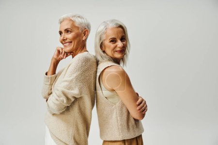 stylish senior woman smiling at camera and standing back to back with happy female friend on grey