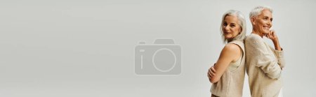 Téléchargez les photos : Modèles de mode séniors enchantés posant dos à dos tout en se tenant sur fond gris, bannière - en image libre de droit