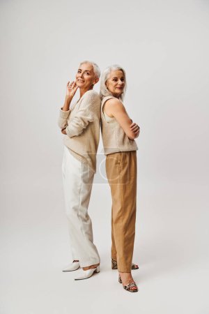 longitud completa de las mujeres ancianas de moda de pie espalda con espalda en gris, amistad y envejecimiento positivo