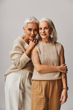 mature woman leaning on shoulder of happy female friend looking at camera on grey, graceful aging