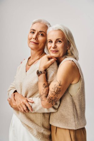 joyous senior woman with silver hair and tattoo embracing fashionable female friend on grey