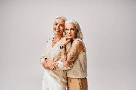 mature tattooed lady hugging cheerful and fashionable female friend on grey, positive aging