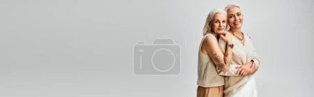 Téléchargez les photos : Insouciant tatoué senior femme étreignant heureux et élégant ami féminin sur fond gris, bannière - en image libre de droit
