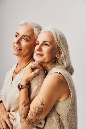 Foto de Mujeres mayores soñadoras y felices en maquillaje y accesorios dorados mirando hacia otro lado en gris, belleza sin edad - Imagen libre de derechos