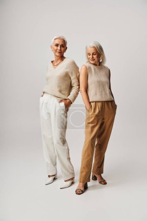 Téléchargez les photos : Positives dames mûres en tenue décontractée élégante posant avec les mains dans les poches sur gris, pleine longueur - en image libre de droit