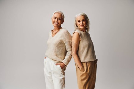 Téléchargez les photos : Femmes âgées souriantes en vêtements à la mode debout dos à dos avec les mains dans les poches sur gris - en image libre de droit