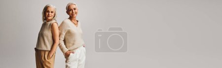 Photo for Happy senior models in trendy clothes posing back to back with hands in pockets on grey, banner - Royalty Free Image