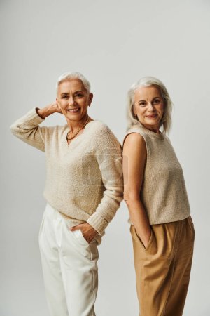 smiling mature female friends standing back to back with hands in pockets on grey, Vanity Fair style