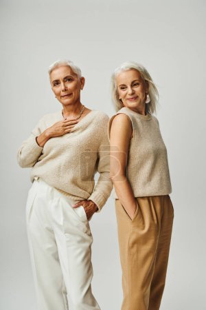 senior ladies in trendy casual attire standing with hands in pockets on grey, lifelong friendship