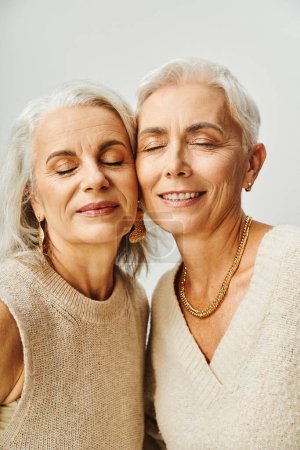 joyeuses femmes âgées en accessoires dorés et maquillage avec les yeux fermés sur gris, amis à vie